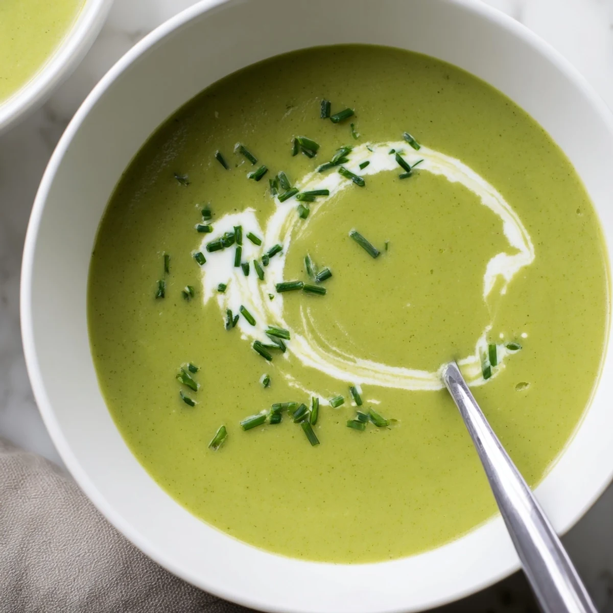 Vibrant green Creamy Broccoli Soup in a white bowl, garnished with fresh chives and a swirl of rich cream, perfect for a cozy dinner.  